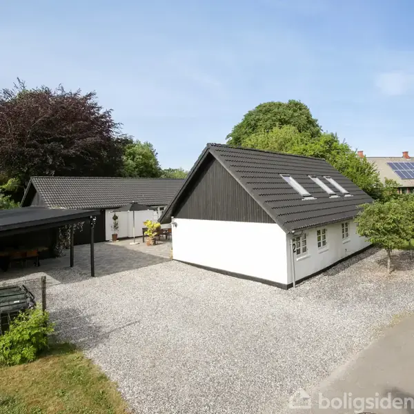 Et hvidt hus med mørkt tag står på en grusbelagt plads. Ved siden af findes en carport og en have. Omgivet af træer på en stille vej.