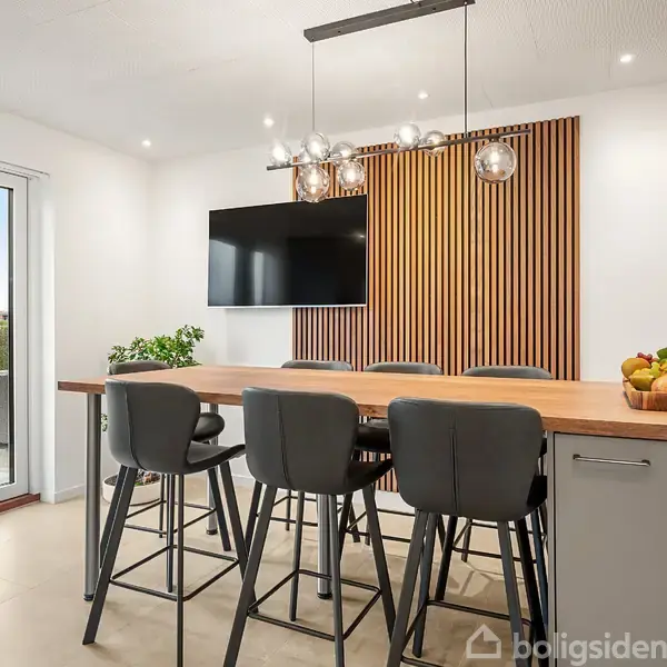 Et spisebord med høje stole står i køkkenet, bagved hænger en fladskærm på en væg dekoreret med træpaneler. Frugtkurv, planter og åbent glasdør til terrasse ses.