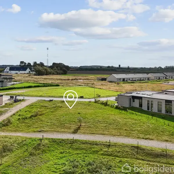Grønt græsareal med placering mærket i midten. Moderne huse ses omkring området, omgivet af flade landskaber og himmel med lette skyer.