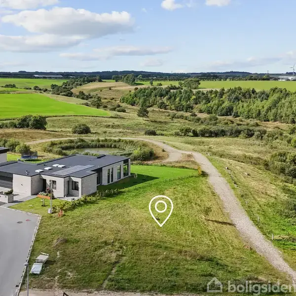 En moderne, fladtaget villa ligger stille blandt grønne marker. Veje og stier omkranser huset, mens åbne landbrugsarealer og skove udgør baggrunden, under en let overskyet himmel.