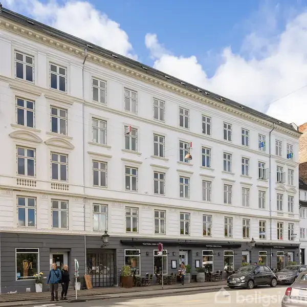 Et fem-etagers, hvidt bygning står langs en byvej. Folk går forbi foran butikker på stueetagen. Luften er klar med spredte skyer over.