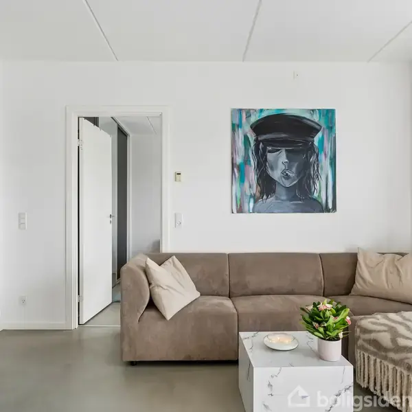 Brun sofa står i en lys stue med hvidmalede vægge. Over sofaen hænger et portrætmaleri. Et hvidt sofabord med en plante og lys dekorerer rummet. En dør er åben til venstre.