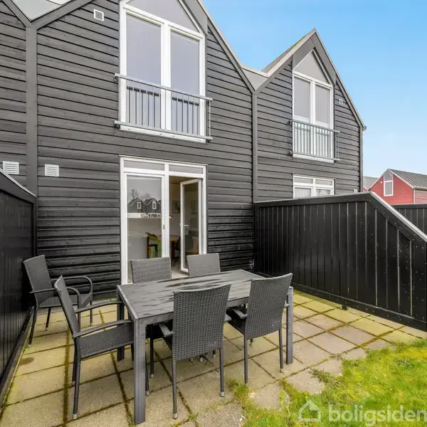 Sort træhus med udendørs terrasse, hvor et sort bord og seks stole står placeret. Terrassen er indrammet af et stakit, og i baggrunden ses en velholdt græsplæne.