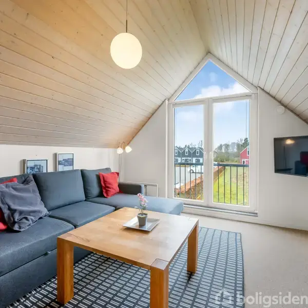Sofa med røde puder står ved et træbord i en stue med skråloft og stort trekantet vindue. TV hænger på væggen, og der er moderne indretning.