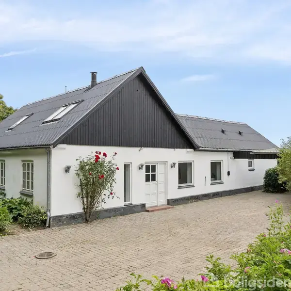 Hvidt hus med sort tag, roser ved indgangen. Står på en brostensbelagt gårdsplads omgivet af grønne buske og træer under en blå himmel.