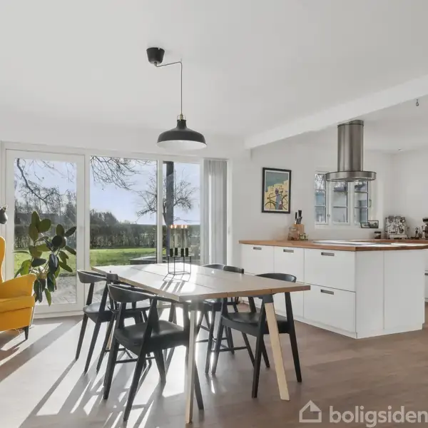 Spisestue med træbord og sorte stole, oplyst af sollys gennem store vinduer. Et gult lænestol står ved siden af en lampe og plante. Moderne køkken i baggrunden.