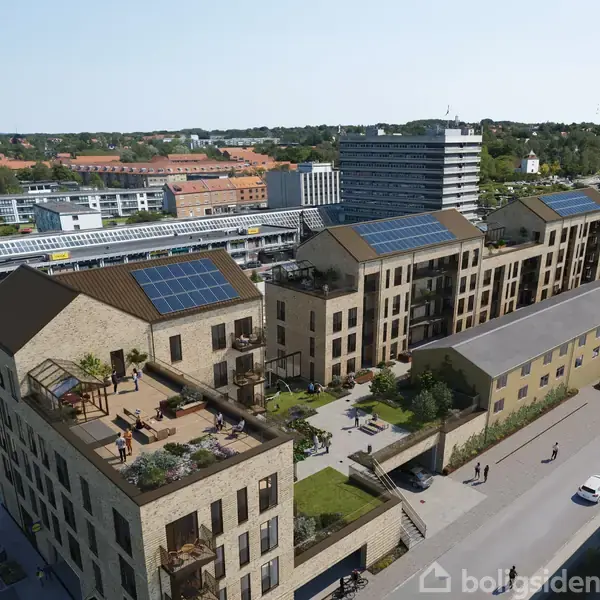 Moderne lejlighedskompleks med solpaneler på tage, placeret i en bydel omgivet af andre bygninger og veje. Mennesker ses gående på fortovet.