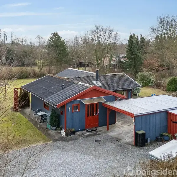 Et blå og rødt træhus ligger stille i en have omkranset af træer. Der er en grusvej foran huset samt en carport på højre side.