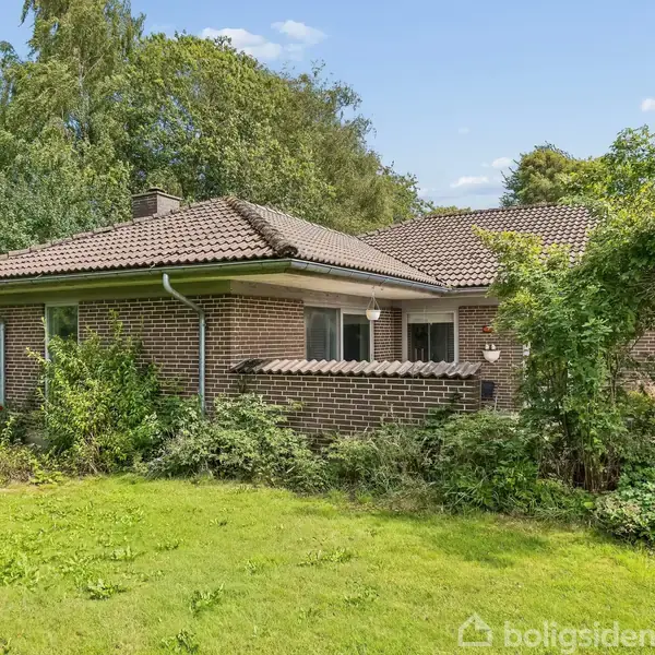 En etplans murstenshus med brun tag, omkranset af frodig vegetation og en velplejet græsplæne. Huset har store vinduer og ligger i en skovrig have med høje træer.