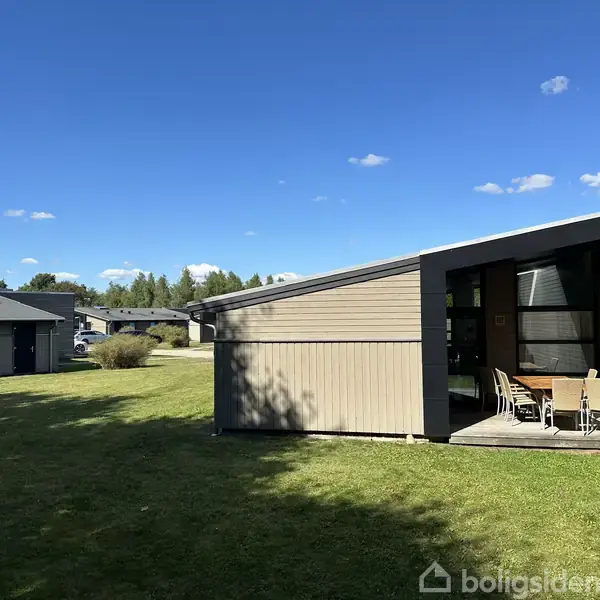 En moderne træbygning med skråt tag står stille i en velplejet græsplæne. Huset har en terrasse med bord og stole. Skove og flere huse ses i baggrunden under en klar blå himmel.