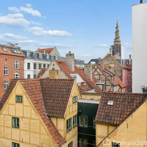 Traditionelle danske byhuse med røde tegltage står tæt sammen. I baggrunden ses kirketårn mod en blå himmel med få skyer, omkranset af moderne bygninger.