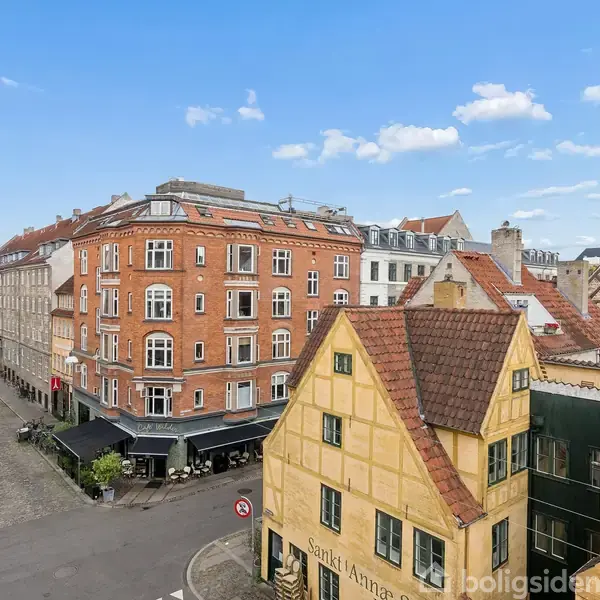 Røde murstensbygninger med hvide vinduer omgiver en brostensbelagt gade. En gul bygning står i forgrunden, mens en kirketårn ses i baggrunden under en blå himmel.