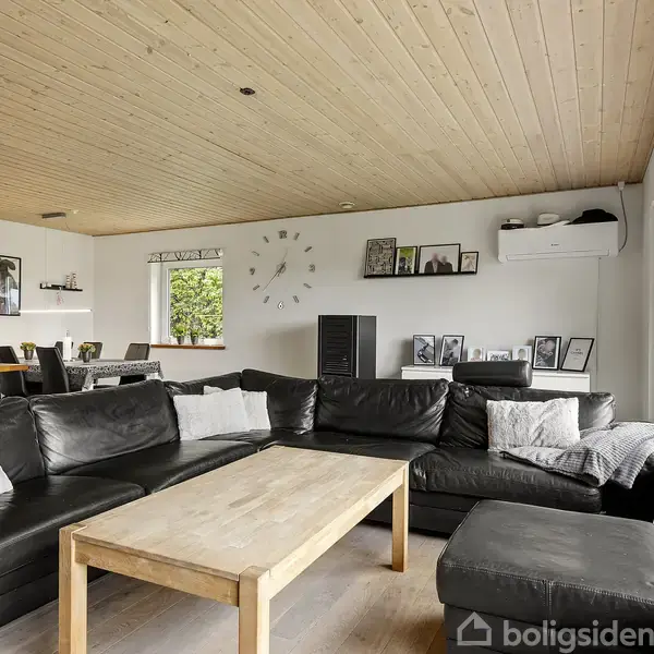 En sort lædersofa står omkring et lyst træbord i en moderne stue. Store vinduer bagved slipper lys ind, og væggen er dekoreret med billeder og en stor vægur.