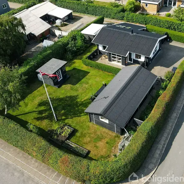 Et sort-hvidt hus med sort tag står ved siden af et mindre udhus på en velplejet græsplæne. Området er omgivet af hække og en vej ses til højre.