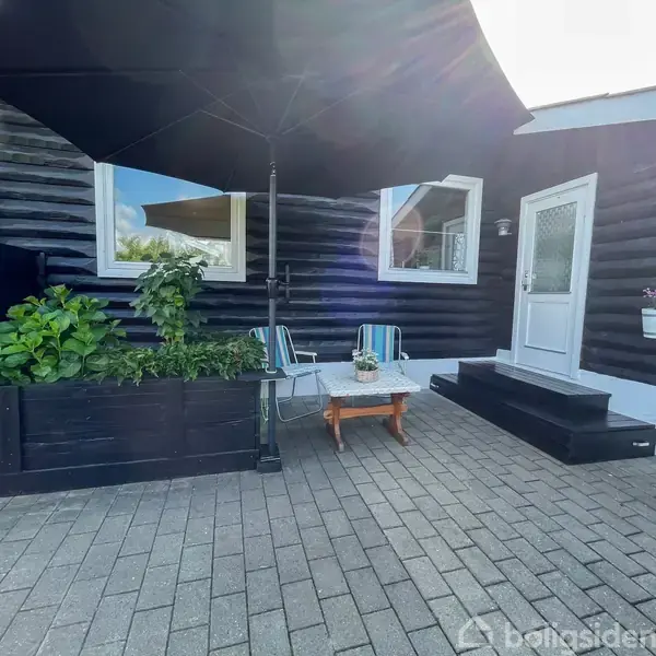 Sortmalet træhus med hvide dør- og vinduesrammer, står ved terrasse med klinkebelægning. To stole og et lille bord med blomster står under en stor sort parasol. Plantekasser med grønne planter findes også.