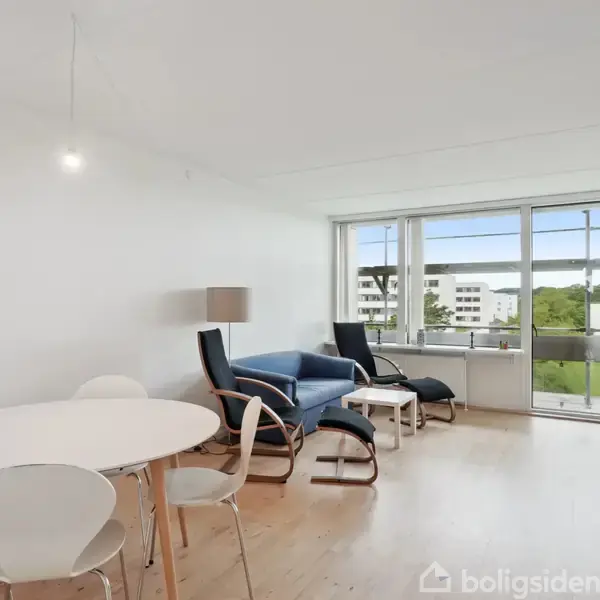 En lys stue med et rundt spisebord og fire stole til venstre. Til højre ses en blå sofa med et lille bord. Store vinduer giver udsigt til grønne områder og bygninger.