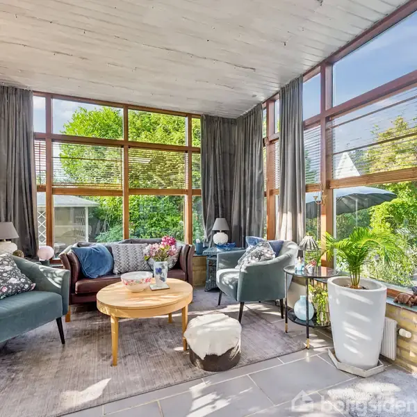 Et lyst værelse med store vinduer viser to lænestole og en sofa omkring et rundt træbord. Blomster, lamper og træer ses gennem vinduerne, hvilket skaber en hyggelig atmosfære.