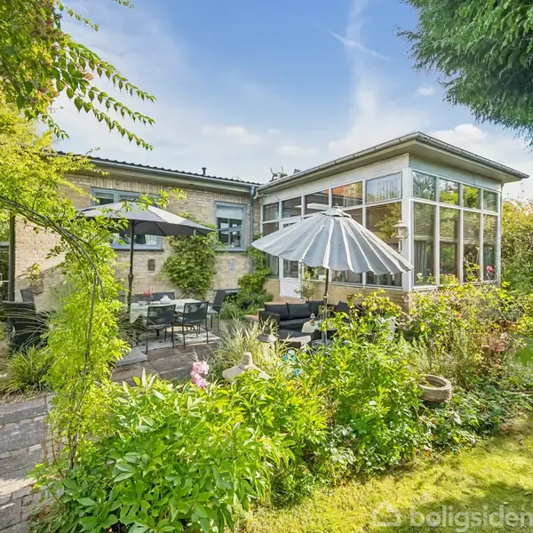 Et hus med store vinduer ud mod en frodig have. En terrasse er udstyret med hvide og sorte parasoller samt havemøbler. Grønt løv omgiver området.