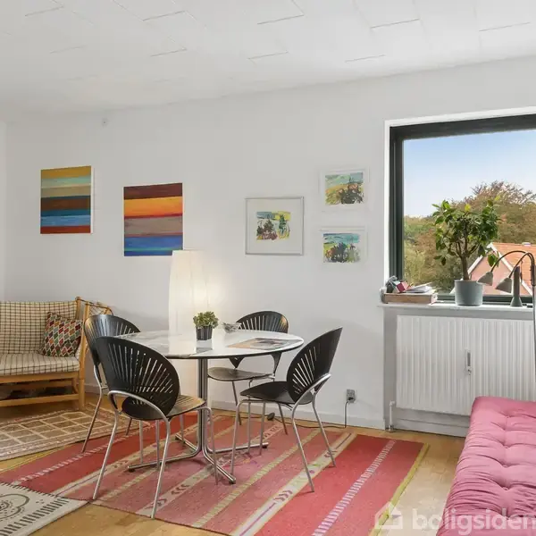 Spisebord med fire stole; lamperne lyser. Omgivet af sofaer og farverige kunstværker på hvide vægge. Et vindue giver udsigt til naturen udenfor. Tæpper dækker trægulve.