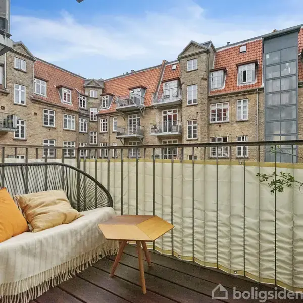En balkon med en sort kurvestol fyldt med puder og tæppe, står ude med et lille træbord. Det omgivende miljø er en klassisk murstensbygning med røde teglstenstag.