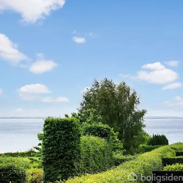 Røde tegltage og grønne hække strækker sig mod en klar blå himmel med skyer. I baggrunden ses en stor træ og en rolig vandoverflade.