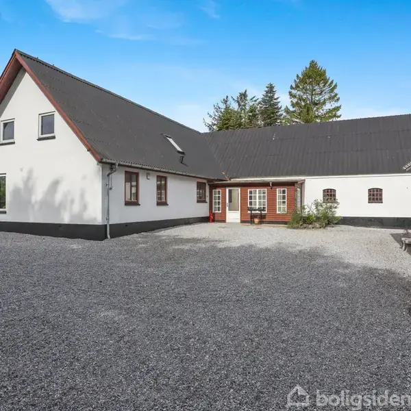Et hvidt, moderne hus med sort tag står på en grusbelagt gårdsplads, omgivet af træer. Huskonstruktionen er vinklet og har flere vinduer og en rød, dobbeltdør.