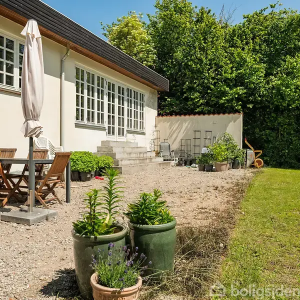 Et hvidt hus med store vinduer foran en terrasse med et bord og seks stole. Parasol står ved siden af, omgivet af potteplanter. Græsplæne og buske i baggrunden.