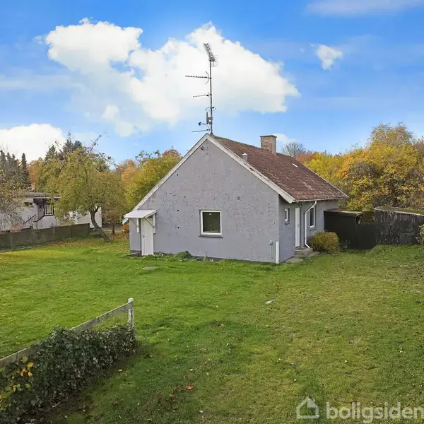 Et lille gråt hus står stille i en rummelig, grøn have omgivet af træer og buske under en blå himmel med skyer.