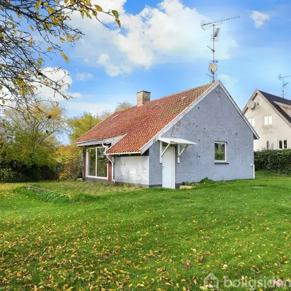 Et hvidt hus med rødt tag står på en grøn græsplæne omgivet af træer. I baggrunden kan man se en større bygning. Himlen er delvist skyet.