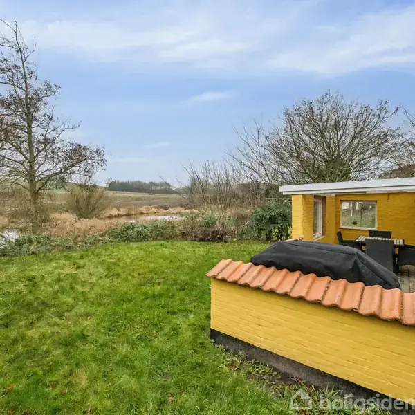 Et gult murstenshus med terrasse er omgivet af grøn have med udsigt til en eng og træer. En grill er dækket på terrassen, og landskabet er åbent og naturligt.