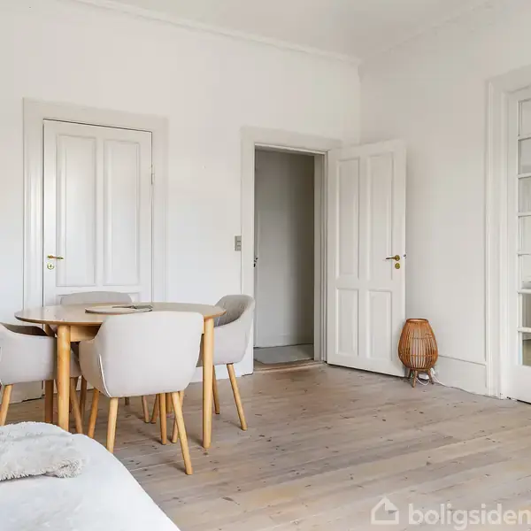 Et rundt træbord med beige stole står i en lys stue med trægulv; to lukkede døre og en åben dør i baggrunden, og en plante ses til venstre.