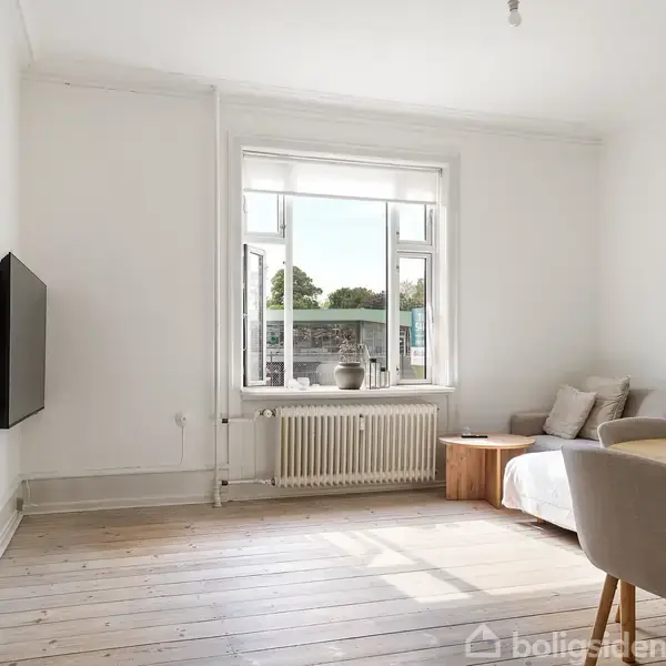 Stue med moderne indretning; en sofa står langs væggen, et tv er monteret på væggen, og et bord er placeret ved siden af en stol. En stor vindue lader dagslys strømme ind.