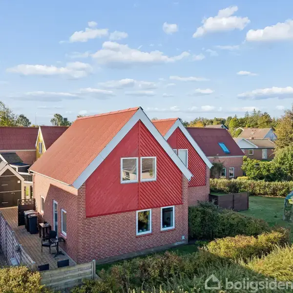 Et rødt murstenshus med en rød gavl og tag står stille. I baggrunden er der flere huse og grønne områder under en blå himmel med skyer.
