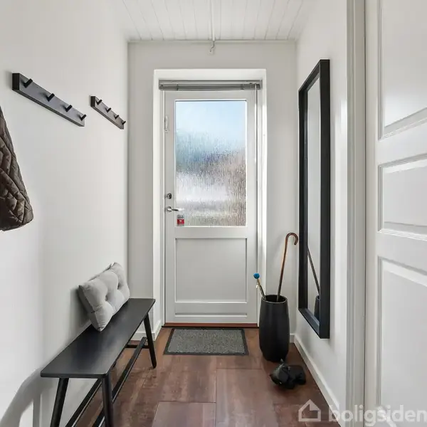 En entré har en bænk med pude til venstre, knagerække med jakker, og en dør for enden. En paraply står ved siden af døren. En lille måtte ligger på gulvet.