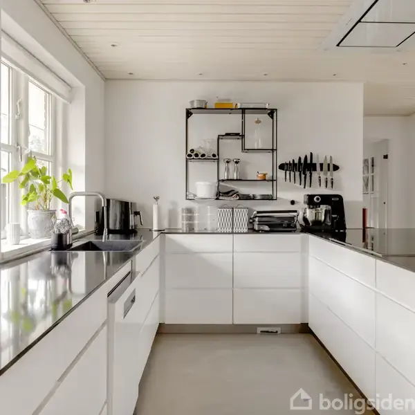 Moderne køkken med hvide skabe, sort bordplade og stål-apparater. Kaffemaskine står på bordet. Indendørs planter ved vinduet. Vægmonterede hylder med glas og kopper.