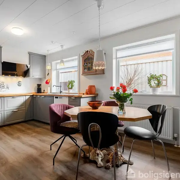 Et moderne køkken med grå skabe og træbordplade. Rundt bord omgivet af forskellige stole i centrum, pyntet med blomster. Vinduet har udsigt til en bygning. Skilt på væggen: "De vigtigste ting i livet er ikke ting...".