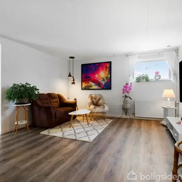 Stue med brun sofa og hvidt sofabord på tæppe, hængende lampe ovenfor. Vækst i hjørnet, vindue med lilla blomst. TV på væggen. Lys trægulv. Kunstnerisk maleri på væggen.