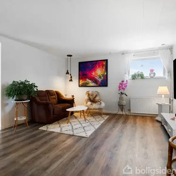 Brunt sofaarrangement med sofabord, placeret på et tæppe. Indrammet kunstværk på væggen. Vindue med planter og lysindfald. Trægulv, rum oplyst af bordlamper.