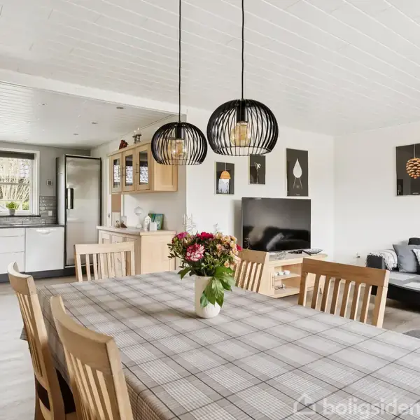Spisebord med ternet dug står i centrum, omgivet af træstole og to hængende lamper. Køkken i baggrunden med hvidt interiør. Sofa og vægdekorationer er synlige i stuen.