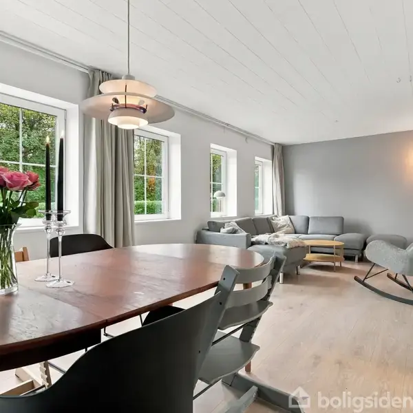 Rundt spisebord med vase og lyseholder står i lys stue med grå sofa, lænestol og store vinduer. Hvide lofter og vægge skaber en lys atmosfære.