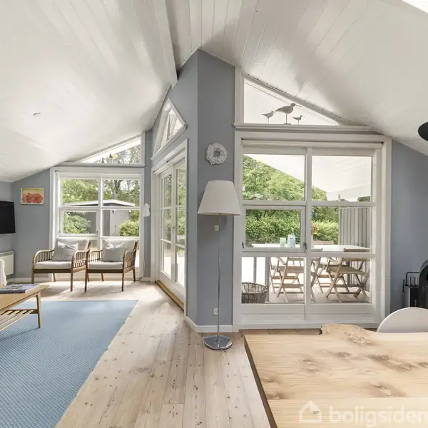 Stue med store vinduer fyldt med naturligt lys. En brændeovn står ved siden af spisebordet i træ, mens sofaen og stolene er placeret på et blåt tæppe. Udsigt til terrasse.