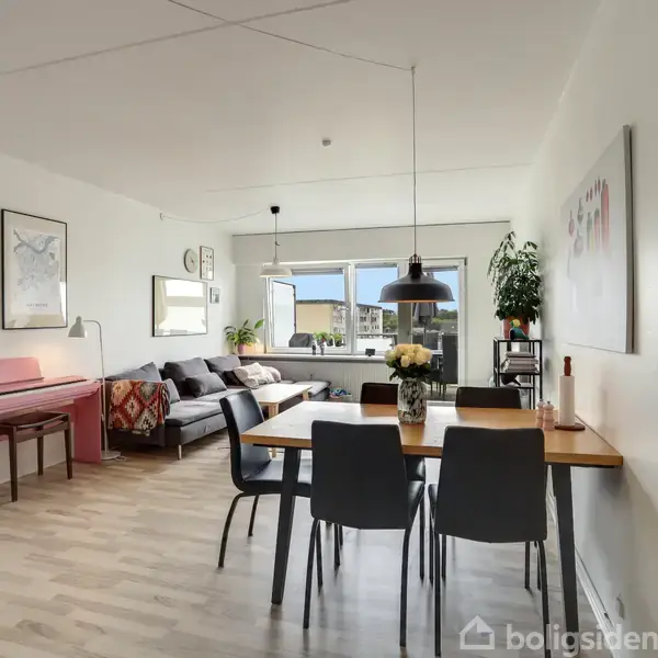 Spisebord med seks stole står i en lys stue med parketgulv. Til venstre ses et lyserødt klaver og sofa, mens en stor plante står nær vinduerne. Billeder hænger på væggene.