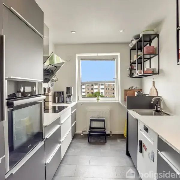 Et moderne køkken med grå skabe og hvide bordplader. Det er veludstyret med ovn, kaffemaskine og vask. Vinduet viser en udsigt til boligblokke udenfor. Hylder bærer køkkenartikler.