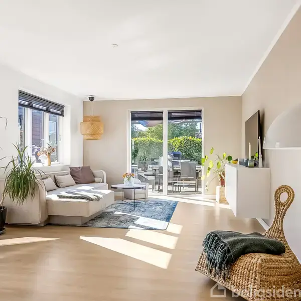 Stuen har en stor, lysegrå sofa med puder, placeret i en lys og rummelig stue. Et rundt sofabord står foran sofaen. Store vinduer og en dør leder til en terrasse.