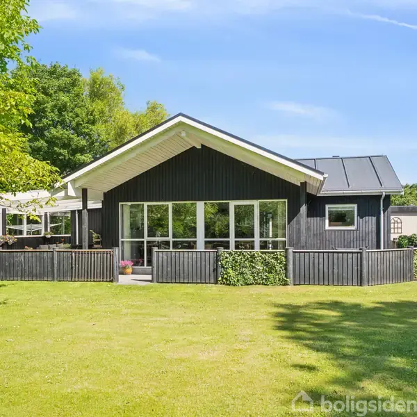 Sort hus med store vinduer beliggende i grøn have med træer. Huset har en veranda og er omgivet af græsplæne og buske, under en klar blå himmel.