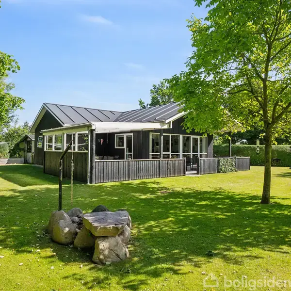 Et sort træhus står stille på en velplejet græsplæne omgivet af grønne træer og buske. En stensætning ses i forgrunden, og himlen er klar og blå.