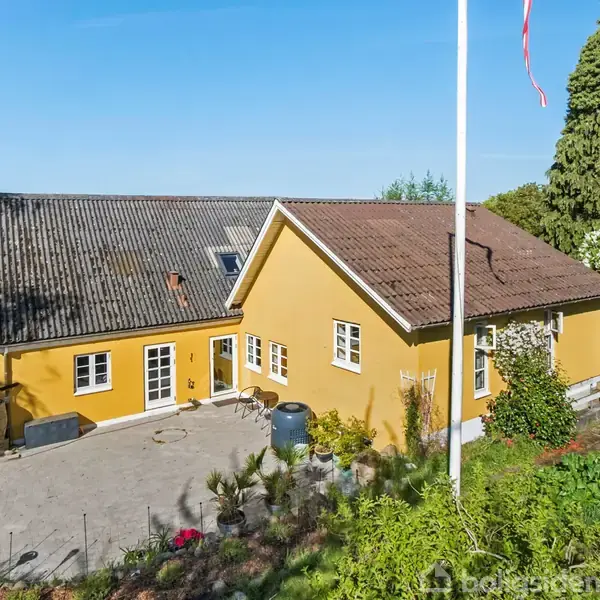 En gulmalet husbygning står stille i en haveomgivelser med grøn vegetation og en flagstang. Blå himmel og marker ses i baggrunden.