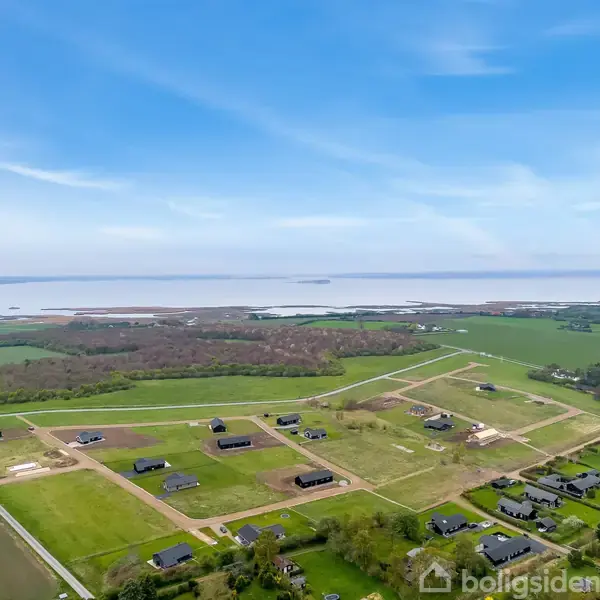 Landsbyhuse ligger på spredte grunde, omgivet af grønne marker og fladt landskab. Havet ses i baggrunden under en klar blå himmel, hvilket skaber en rolig atmosfære.