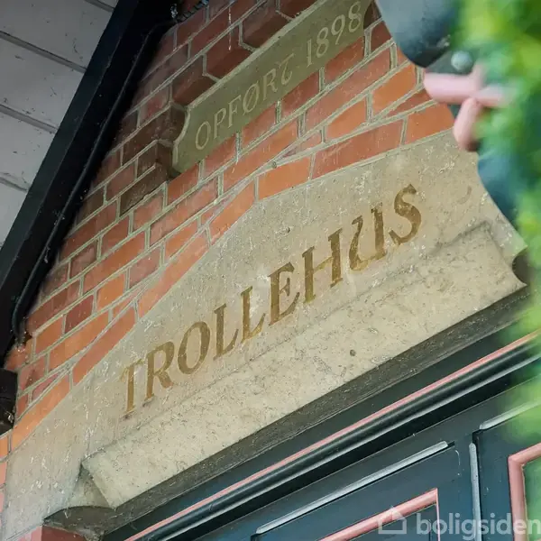 En sten indgraveret med teksten "TROLLEHUS" er over en dør, omkranset af røde mursten. Ovenfor ses "OPFØRT 1898" på en mindre sten.