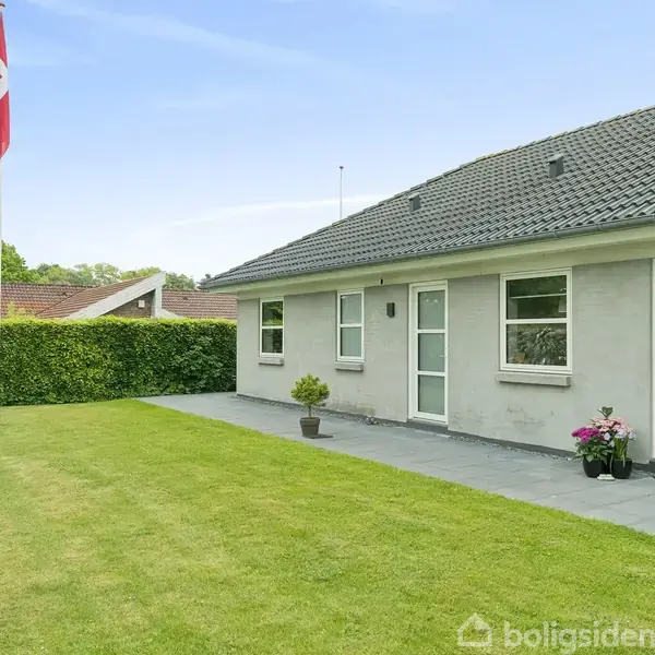 Hus står stille i en velplejet have, omgivet af hække og træer. Trampolin og terrasse med møbler samt grill ses. Flagstang står til højre i billedet.
